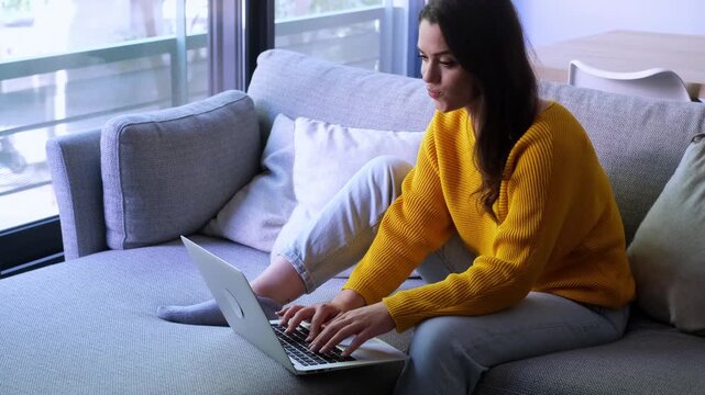 Young professional woman sitting on a comfortable couch in her modern living room, working remotely on her laptop computer, focused on typing an important message or email