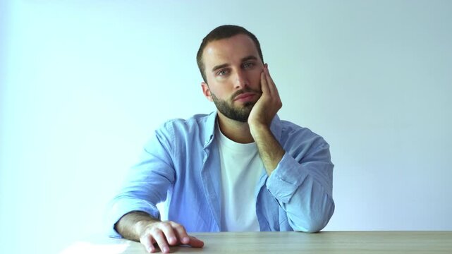 Young handsome man feeling bored and tired while sitting at a desk and waiting for something, his expression showing disinterest and impatience on a clean white background