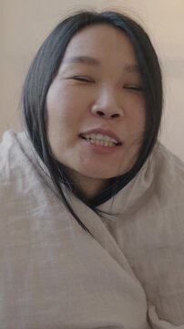 Slow motion portrait of adorable Asian lady looking at camera from under blanket lying in bed sending kiss smiling looking at camera in bedroom at home