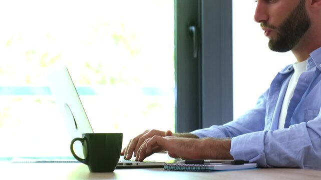 Exhausted young freelancer feeling neck ache and muscle strain while typing on a laptop computer keyboard at his desk, highlighting the health issues of a sedentary office lifestyle