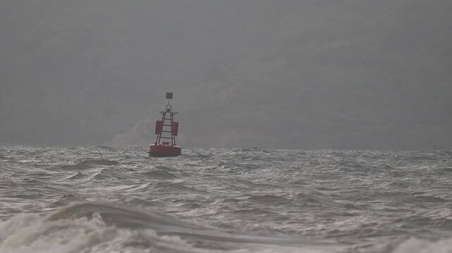 A red channel marker navigation buoy floating alone and bobbing on rough wavy gray sea water with choppy waves during overcast misty rainy day at Lembar Bay, Lembar Village, West Lombok Regency.