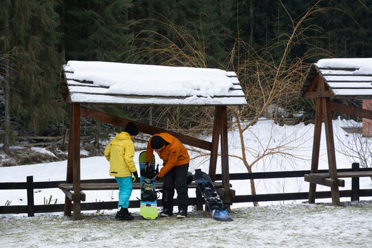 Two friends prepare for a snowboarding adventure in a winter wonderland near a tranquil forest, surrounded by fresh snow and wooden shelters in the early morning light