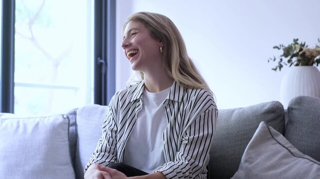 Beautiful young woman sitting on a modern sofa in her living room, laughing sincerely and showing genuine emotion, enjoying a pleasant and amusing conversation with someone off-camera