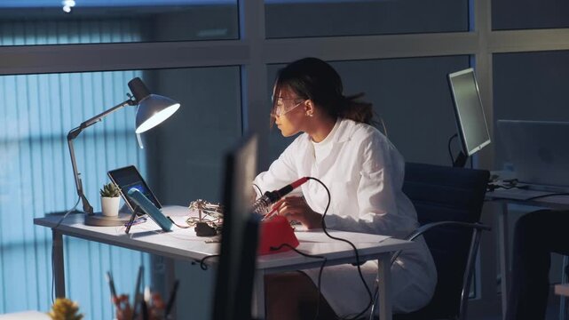 African American scientist wearing white coat and protective glasses testing a circuit board with soldering iron and multimeter in a bright laboratory workspace.