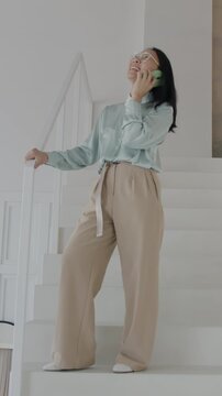Cheerful Asian woman talking on mobile phone standing on stairs in luxurious house. Smartphone communication and stylish interior concept.