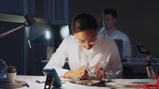 An African American female engineer wearing protective glasses tests a circuit board with a multimeter in a technical electronics laboratory with a male colleague present.