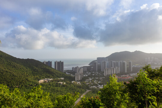 Panoramic high-angle view of Sanya city, Hainan, China. Modern skyscrapers between tropical mountains and coastline under dramatic cloudy sky. Travel, urban growth of popular Chinese seaside resort.