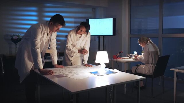 Multiracial electronics engineers in white lab coats collaborate on control schematics around a large table in a modern laboratory with technical equipment and natural light.