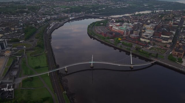 Aerial view of Peace Bridge spanning the River Foyle with city buildings and sunset light in Derry, Northern Ireland, United Kingdom.