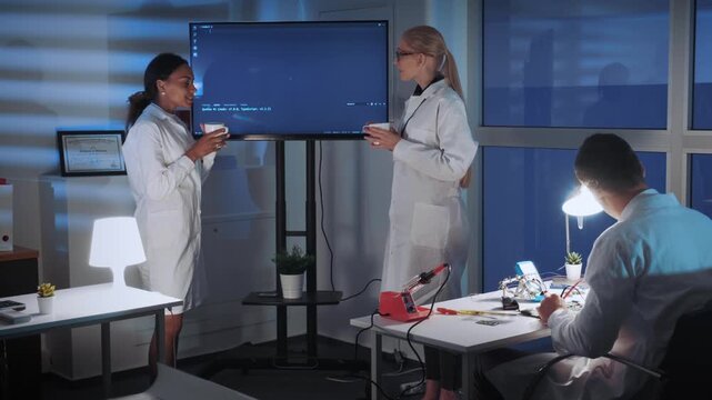 Two female engineers in lab coats discuss technical data near a large monitor while a male colleague works with equipment in a bright modern laboratory.
