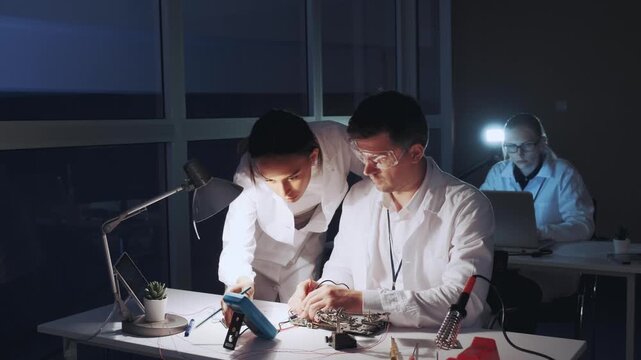 Four electronics engineers in white lab coats work on a motherboard using a multimeter and soldering tools in a modern laboratory at nighttime.
