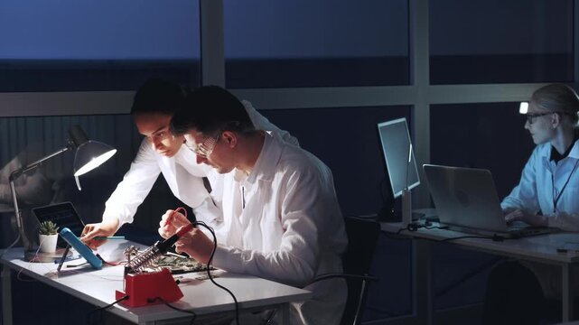 Two engineers test a circuit board with multimeter and soldering tools while two others program laptops in a nighttime electronics laboratory.