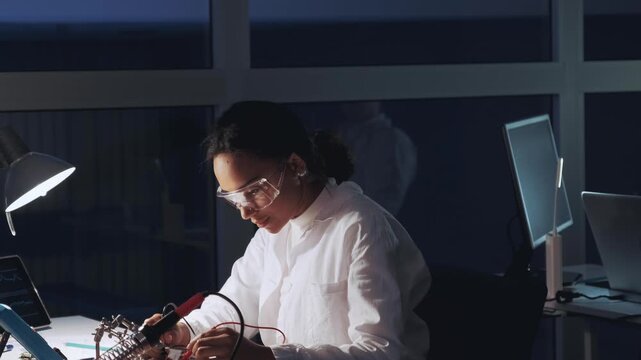 African American female engineer wearing protective glasses works on soldering and testing circuit boards in a high-tech laboratory at nighttime, celebrating success.