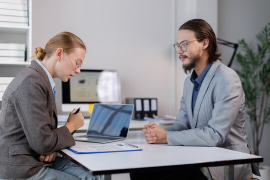Recruiter holding a pen and looking at a document, interviewing a male candidate across a desk in a bright office environment, discussing a job application