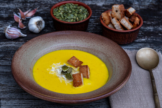 Pumpkin cream soup with croutons and seeds in ceramic bowl. Delicious yellow squash puree soup served with crunchy croutons pumpkin seeds and garlic on table, autumn comfort food background