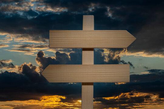 Blank wooden signpost with arrows pointing opposite directions at sunset, closeup. Empty wooden crossroads sign pointing left and right against dramatic sunset sky with dark clouds