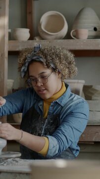 Female potter working with throwing wheel in workshop while man doing inventory checking ceramic pots on shelves. Small business and creative start-up concept.