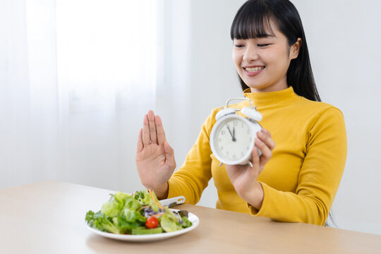 Smiling woman holding alarm clock and making stop gesture before eating salad, wellness concept for meal timing, mindful eating, healthy routines, nutrition and balanced lifestyle habits.