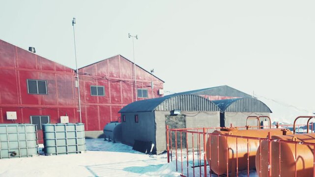 People are working at a research station surrounded by snow. There are red and gray buildings, large tanks, and a clear sky above them, showing the cold environment of the setting.