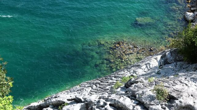 Clear turquoise sea water moving along a rocky coastline with visible stones under the surface on a sunny day.