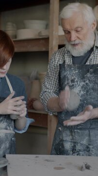 Senior man teaching grandson to work with clay talking enjoying leisure time in pottery workshop. Family business and relationship concept.