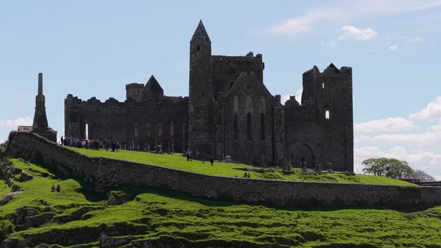 Aerial view of the historic Rock of Cashel, a medieval fortress set on a grassy hill under a blue sky with scattered clouds in Cashel, County Tipperary, Ireland.