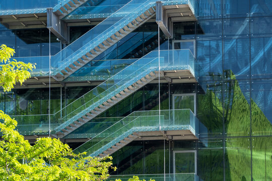 Glass building exterior with staircases and green reflections combines modern architecture with urban vegetation across a layered transparent facade