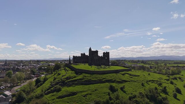 Aerial view of the historic Rock of Cashel stone fortress atop a lush green hill under a bright blue sky in Cashel, County Tipperary, Ireland.
