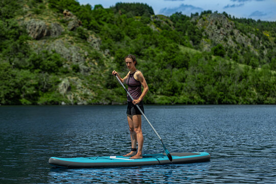 Athletic slim female stands on paddleboard and paddles on blue water lake or river with trees and cliffs on shoreline, wearing shorts tankini, enjoying outdoors exercise in sunshine in summer holiday