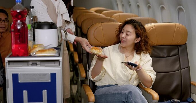 Female flight attendant hands packaged bread to adult Asian woman seated with smartphone inside modern aircraft cabin, representing inflight service, airline hospitality and passenger experience.