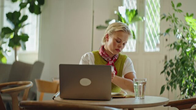 Woman works on a laptop and writes notes at a cafe table, showing remote work, freelance productivity, and studying in a bright indoor workspace with a focused, independent routine.