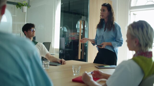 Woman leads a business meeting with her team in a bright office conference room, presenting ideas and guiding discussion around the table in a collaborative workplace.