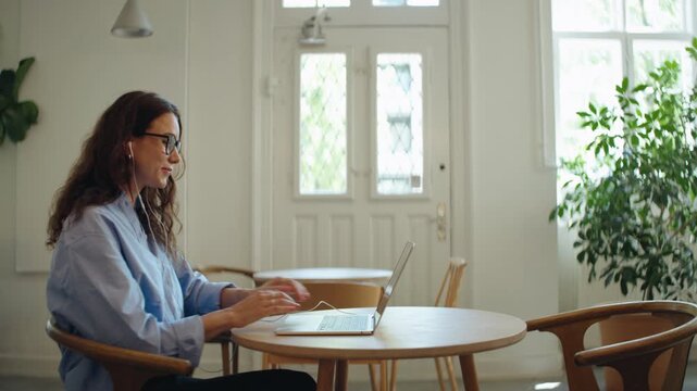 Woman speaks on a video call on her laptop in a bright indoor workspace, wearing earphones and gesturing during an online meeting. Remote work, communication, and freelance business are clearly repres