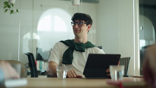 Man speaks and smiles during a business meeting in a bright office, showing work communication, engagement, and presence in a modern workplace discussion while using a tablet at the table.