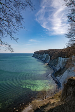 View of the iconic white chalk cliffs of Rugen rising above the turquoise Baltic Sea, framed by forest branches under a clear blue sky in Mecklenburg-Vorpommern, Germany.