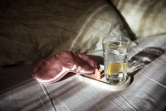 Glass of water with lemon slice and pink sleep mask on a bedside table, soft bedding and natural light creating a calming atmosphere in a bedroom setting