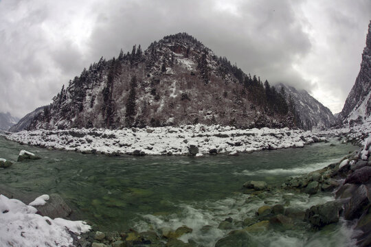 View of the Ushuroi Valley in winter with a flowing river, snow-covered banks, and pine-clad mountains under a cloudy sky in Kalam, Khyber Pakhtunkhwa, Pakistan.