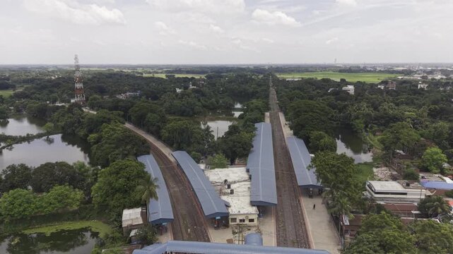 Backward-moving drone footage reveals Poradah Junction railway station in Kushtia, Bangladesh, showing multiple railway tracks, covered platforms, a pedestrian footbridge, and lush green surroundings.