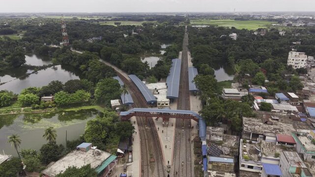 Ascending drone footage gradually reveals Poradah Junction railway station in Kushtia, Bangladesh, with multiple railway tracks, covered platforms, a pedestrian footbridge, and surrounding greenery.