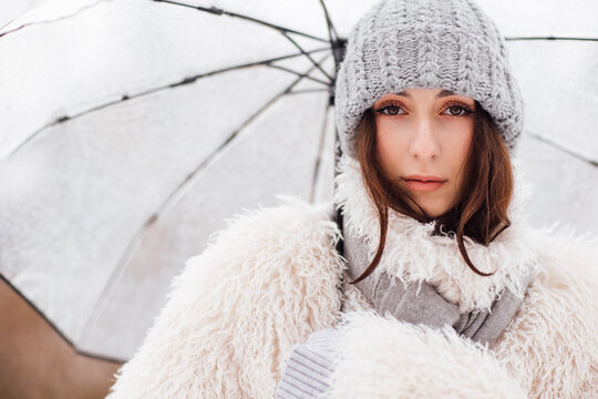 Young woman with long brown hair wearing a cozy white fur coat and gray knitted hat holds a transparent umbrella against a blurred winter background
