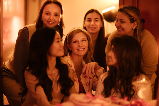 Group of six women of diverse ethnic backgrounds smiling and posing together in a warmly lit indoor setting, with soft colors and a cozy atmosphere
