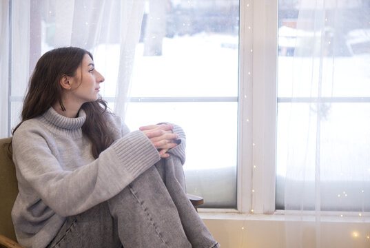 Young woman with long brown hair wearing a gray sweater sits by a window, gazing outside at a snowy landscape with soft natural light illuminating the room