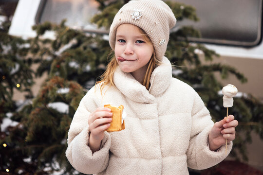 Young girl with blonde hair wearing a beige coat and knitted hat enjoys a snack outdoors, holding a marshmallow on a stick and a pastry in front of evergreen trees