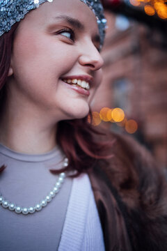 Young girl with red hair wearing a silver headband and pearl necklace smiles while standing outdoors with blurred festive lights in the background