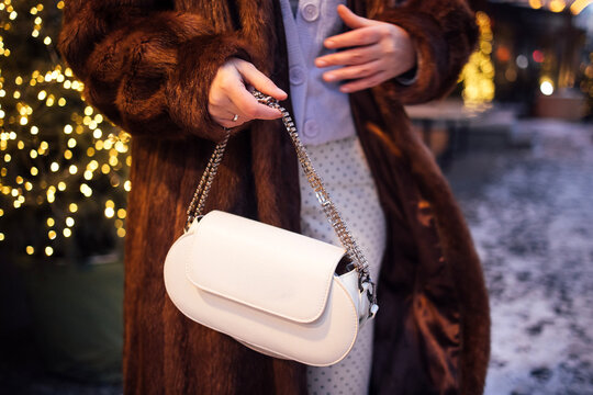 Female fashion model wearing a brown fur coat holds a white handbag while standing outdoors with blurred festive lights in the background during winter season