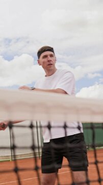 Vertical video of Caucasian young adult man hitting tennis balls with racket over net on clay court in slow motion. Footage suits sports marketing, workout apps, and tennis coaching promos