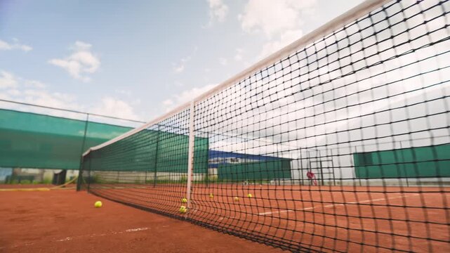 Yellow tennis balls scattered along net on outdoor clay court in slow motion as players train. Clip serves sports marketing, tournament promos, tennis coaching campaigns