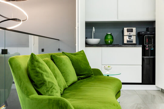 Green velvet sofa with plush cushions is placed in a contemporary kitchen featuring white cabinetry, a coffee maker, and a decorative bowl, creating a stylish and inviting atmosphere