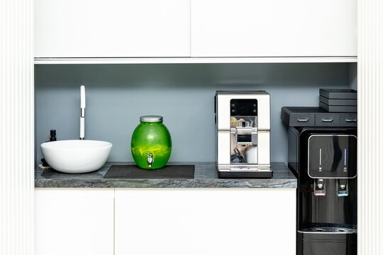 Modern kitchen interior featuring a sleek coffee machine, a green decorative jar, and a stylish water dispenser on a marble countertop against a minimalist backdrop