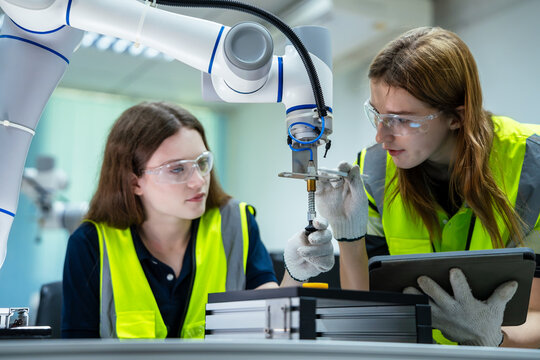 Close up of professional female engineers evaluating and adjusting a robot arm with a digital tablet in a high-tech manufacturing lab. Industrial automation and machine learning concept.